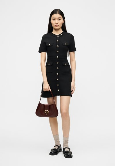 Νέα γυναίκα με μαύρο φόρεμα με κουμπιά κρατά καφέ σκούρη τσάντα, φορά μπεζ κάλτσες και μαύρα μοκασίνια, στέκεται μπροστά σε απλό φόντο.