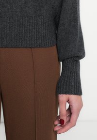 Σκούρο γκρι πλεκτό πουλόβερ με ριμπ μανσέτες, σε συνδυασμό με καφέ υφασμάτινο παντελόνι με υφή. Κοντινή λήψη του μανικιού και του χεριού, παρουσιάζοντας πλούσια αντίθεση χρωμάτων.