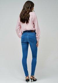 Jeans de mezclilla azul delgados con una cintura de media altura, que cuentan con bolsillos traseros, costuras sutiles y un parche de marca de imitación cuero en la cintura.