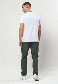 Mand iført en ensfarvet hvid T-shirt, mørkegrønne cargobukser og hvide sneakers, stående med ryggen mod en lys baggrund.