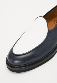Navy leather slip-on shoe featuring a white textured toe cap, round shape, contrasting stitching, and a black rubber sole.