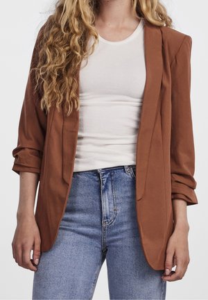 Frau trägt ein braunes Sakko über einem weißen, gerippten Tanktop und hochtaillierte hellblaue Jeans, steht vor einem einfarbigen hellen Hintergrund.