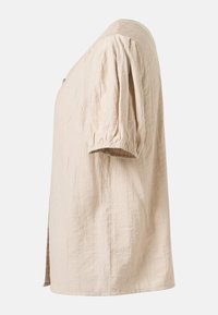 Blouse beige à manches courtes avec manches bouffantes et tissu légèrement texturé, vue de profil sur un fond uni.