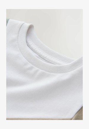 Fehér póló texturált, kerek nyakkivágással, zöld és bézs szövetbetétekkel a váll közelében. Simább, könnyű pamut anyagból.