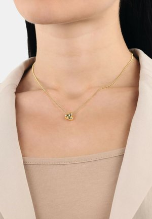 Collar de oro con un colgante de nudo retorcido, sobre una cadena delicada, lucido sobre una blusa de tono neutro, que destaca su textura pulida.