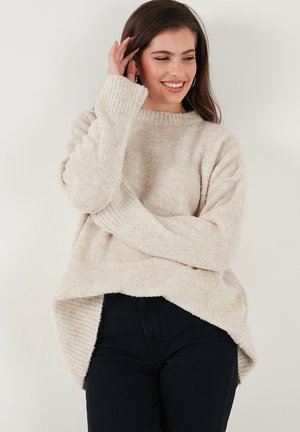 Pull en crème flou avec un col et des poignets côtelés, offrant une coupe décontractée et des fentes sur le bas. Associé à un jean sombre.
