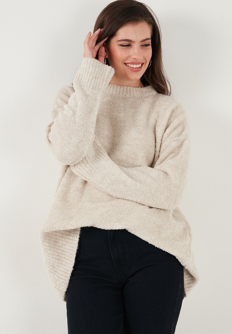 Maglione fuzzy color crema con scollatura e polsini a coste, caratterizzato da una vestibilità comoda e spacchi laterali all'orlo. Abbinato a jeans scuri.