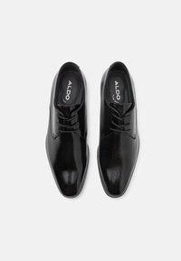 Pantofi de dress din piele neagră cu textură netedă, vârf ascuțit și design cu șireturi. Prezintă logo pe tălpi și o strălucire subtilă.
