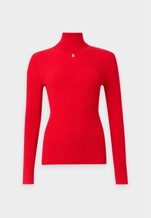 Femme aux cheveux bouclés portant un pull col roulé rouge ajusté, un pantalon large marron à fines rayures et des talons ouverts noirs, debout les mains dans les poches.