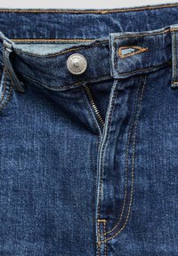 Jean en denim avec braguette partiellement ouverte, bouton en métal visible et coutures orange sur tissu bleu foncé.