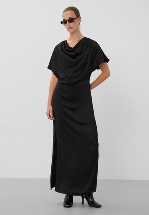 Femme portant une longue robe noire drapée, des bottines noires et de petites lunettes de soleil noires, debout devant un fond blanc uni.