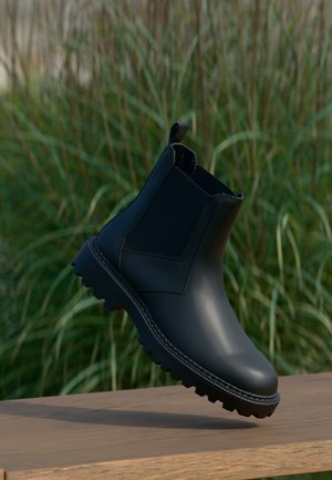 Bottine en cuir noir avec tirant et panneaux latéraux élastiques flottant au-dessus d'une surface en bois, herbe verte floue en arrière-plan.