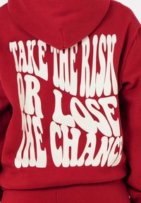 Rode hoodie met witte, golvende tekst "NEEM HET RISICO OF VERLOSL DE KANS" op de achterkant. Zacht fleece materiaal en verstelbare capuchon.
