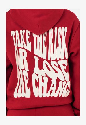 Rode hoodie met witte, golvende tekst "NEEM HET RISICO OF VERLOSL DE KANS" op de achterkant. Zacht fleece materiaal en verstelbare capuchon.