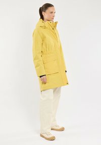 Veste imperméable jaune avec capuche, taille ajustable, deux poches avant et accents noirs. Portée avec un pantalon clair et des chaussures beiges.