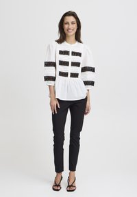Chemisier blanc avec des accents en dentelle noire, manches ballons, fermeture à boutons, ourlet peplum, associé à un pantalon noir ajusté et des talons noirs à lanières.