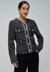 Veste en tweed noir avec un col rond, deux poches avant et des accents de boutons dorés. Les bords effilochés ajoutent de la texture au design.