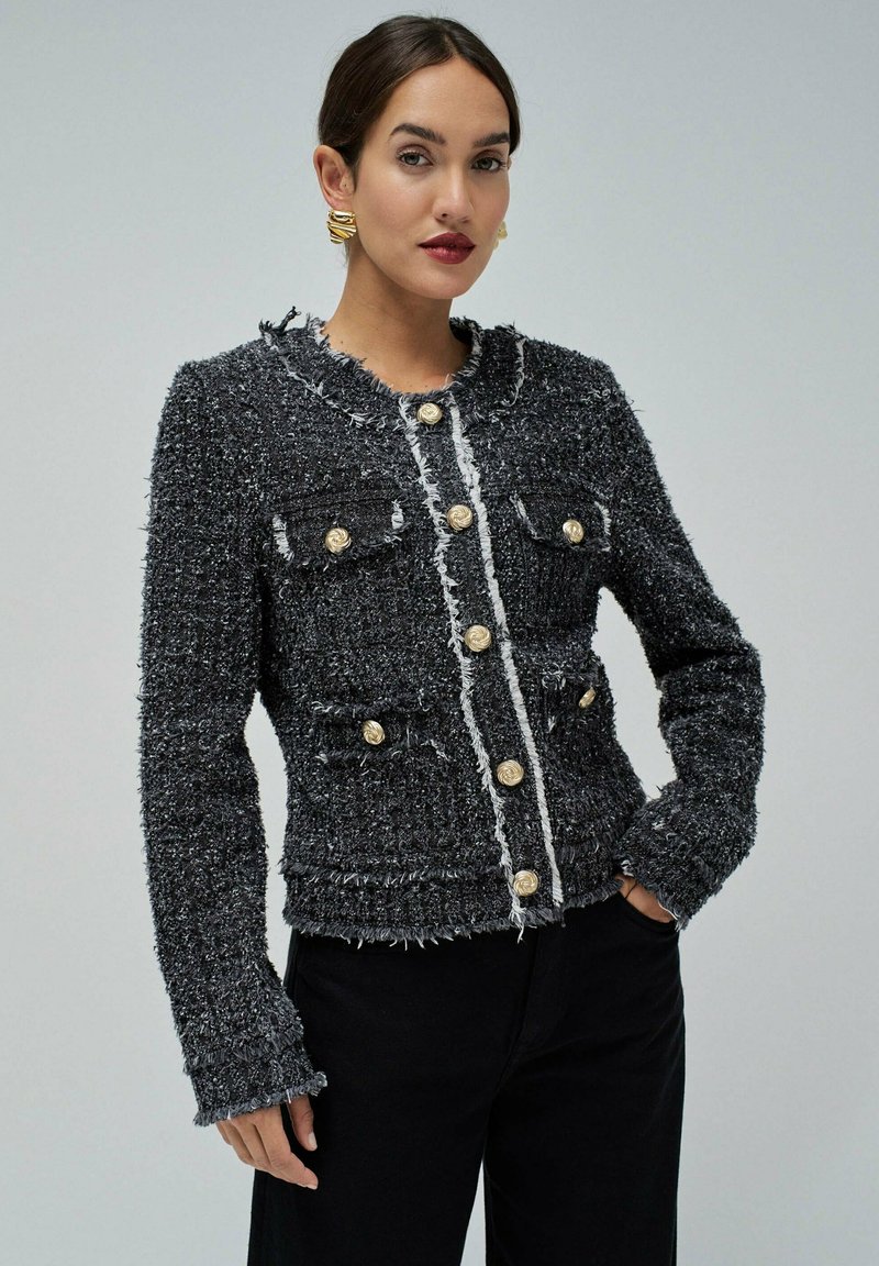 Veste en tweed noir avec un col rond, deux poches avant et des accents de boutons dorés. Les bords effilochés ajoutent de la texture au design.