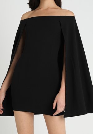 Vrouw draagt een korte zwarte off-shoulder jurk met lange cape-achtige mouwen en zijsplitten die haar armen onthullen.