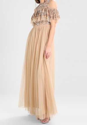 Beige, tot op de vloer vallende jurk met doorschijnende laag, lijfje met geborduurde bloemen en pailletten, ruches aan de cold shoulder mouwen, gedragen met sandalen met bandjes.