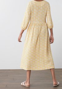 Vestido amarillo con un corsé ajustado, cintura fruncida y mangas abullonadas de tres cuartos. Presenta un sutil patrón geométrico y un dobladillo en capas.