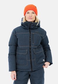 Donna che indossa una giacca imbottita blu scuro con numerose tasche e un berretto di lana arancione, in piedi davanti a uno sfondo neutro.