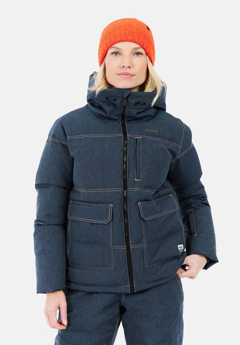 Donna che indossa una giacca imbottita blu scuro con numerose tasche e un berretto di lana arancione, in piedi davanti a uno sfondo neutro.