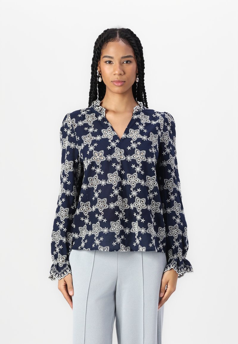 Blusa blu navy con intricati ricami floreali bianchi, caratterizzata da scollatura a V, colletto con volant e maniche lunghe con polsini scallop.
