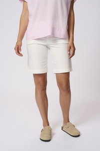 Woman wearing pale pink short-sleeve blouse, white knee-length shorts, and beige slip-on shoes standing against a plain background.
