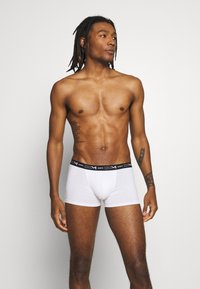 Weiße Boxer Shorts mit einem schwarzen DIM-Bund, mit einer enganliegenden Passform und weichem Stoff, Modell trägt sie, im Profil stehend.