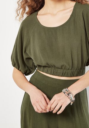 Kvinde iført olivengrøn crop top med puffede ærmer og matchende nederdel, hænderne foldet med perlearmbånd på højre håndled.