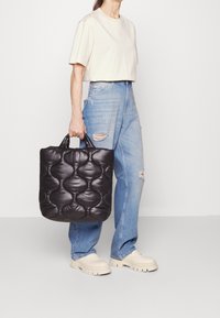 Osoba v béžovém tričku, světle modrých roztrhaných džínách a krémových robustních botách drží černou prošívanou tašku s kulatými rukojeťmi.