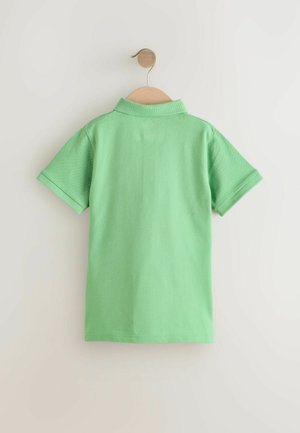 Polo vert suspendu sur un cintre en bois, présentant un col classique et des manches courtes, avec un tissu texturé et sans motifs visibles.