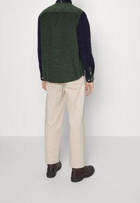 Grön kordfläskeskjorta med marinblå ärmar, knäppta manschetter och rund nederkant, kombinerad med beige byxor och bruna stövlar.