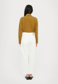 Camicia marrone a maniche lunghe con colletto, abbinata a pantaloni bianchi a gamba larga con tasche posteriori e etichetta. Scarpe nere completano l'outfit.