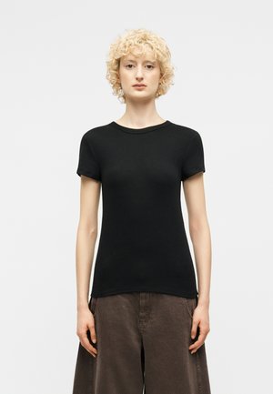 Γυναίκα με κοντά κατσαρά ξανθά μαλλιά, που φοράει εφαρμοστή μαύρη κοντομάνικη μπλούζα και φαρδιά καφέ παντελόνια, στέκεται μπροστά σε απλό φόντο.