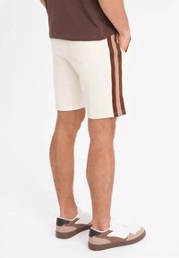 Shorts en crème avec des rayures latérales marron, fabriqués en coton. Accents marron sur des baskets blanches, poches visibles et coupe ajustée.