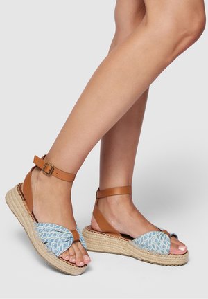 Pies de mujer con sandalias cuña de esparto de patrón azul y tiras de tobillo color canela sobre fondo gris claro.