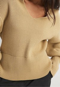 Maglione beige a coste con ampio scollo a V, caratterizzato da una vita aderente e maniche texturizzate, realizzato in un materiale morbido ed elastico.
