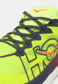 Φωτεινό κίτρινο αθλητικό παπούτσι με μαύρα κορδόνια, μαύρες λεπτομέρειες και κόκκινο λογότυπο στη γλώσσα και στην πλευρά, κοντινή φωτογραφία σε λευκό φόντο.