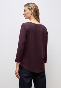 Bordeauxrood langemouwen top met een brede halslijn en een gebogen zoom, gemaakt van zachte stof. Model draagt donkerblauwe jeans, wat een casual uitstraling geeft.