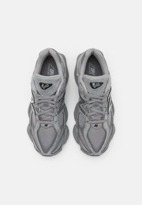 Šedé sieťované a semišové tenisky s robustným dizajnom, ktoré majú textúrovaný zvršok, spevnenú špičku a logo akcenty na jazyku a strane.