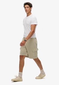 Herenoutfit met een aansluitend wit katoenen T-shirt en knielange beige cargoshorts met zijzakken, gecombineerd met beige sneakers en crew-sokken.