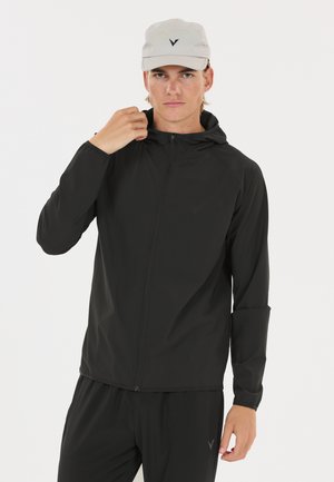 Jeune homme portant une veste à capuche noire zippée et un pantalon noir, ajustant le col de la veste, avec une casquette claire sur la tête.