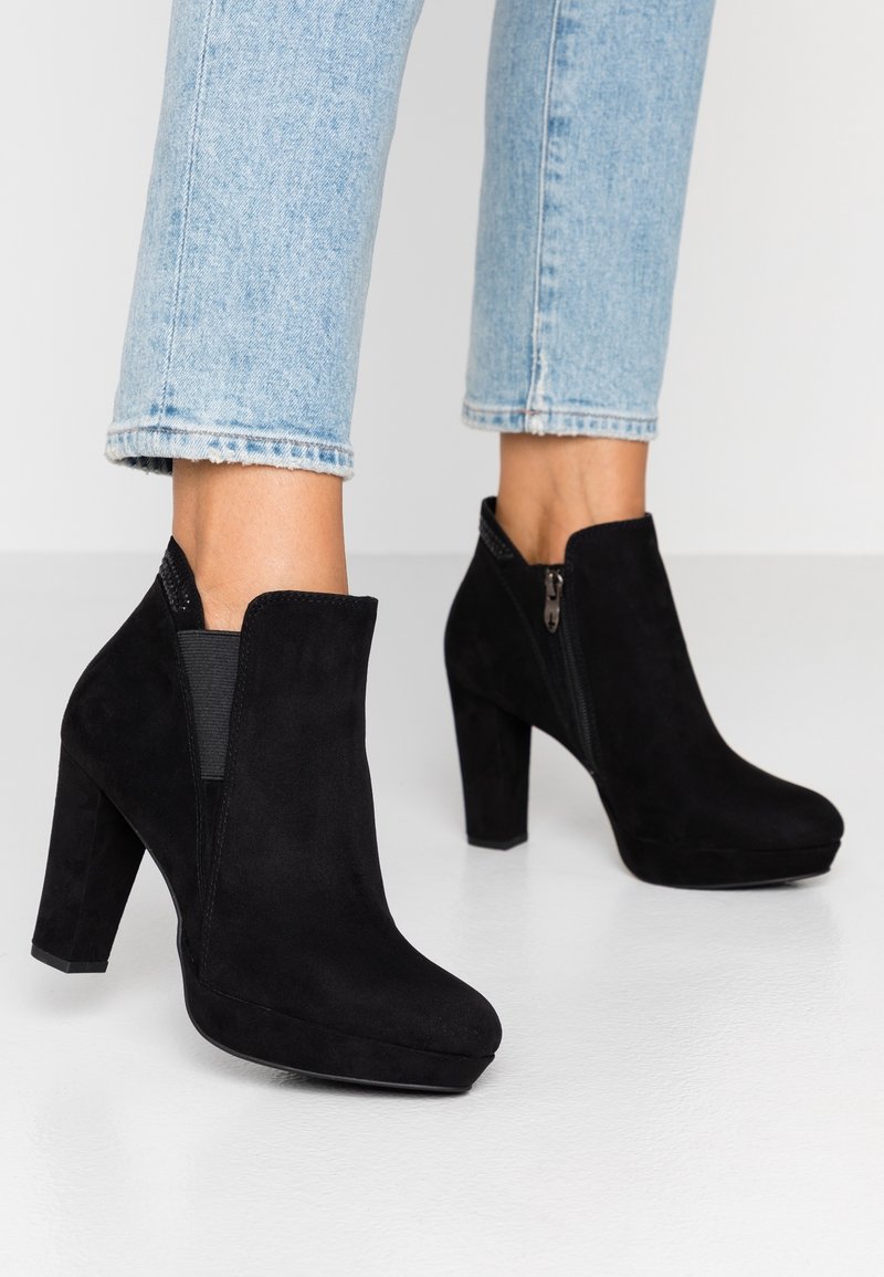 Bottines en daim noir à talons carrés portées avec un jean bleu clair raccourci sur un sol blanc.