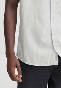 Camisa de lino blanca con botón en el frente, mangas cortas y dobladillo curvado; combinada con pantalones de mezclilla oscuros. Etiqueta visible en el lado.