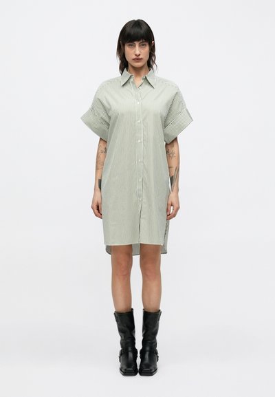 Femme aux cheveux foncés portant une robe chemise à manches courtes à rayures verticales et des bottes noires mi-mollet, debout devant un fond blanc uni.