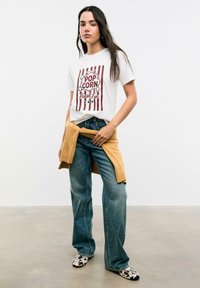 T-shirt graphique blanc avec un design de popcorn rouge, associé à un jean en denim ample et un pull beige noué autour de la taille. Les chaussures ont un motif tacheté.