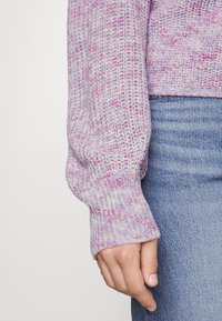 Pull en tricot violet clair avec des accents roses, présentant des poignets côtelés et une coupe ample, associé à un jean en denim.