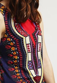 Žena s vlasy po ramena, oblečená v bezramínkových šatech s odvážnými geometrickými a květinovými vzory v červené, modré, žluté, černé a bílé barvě.